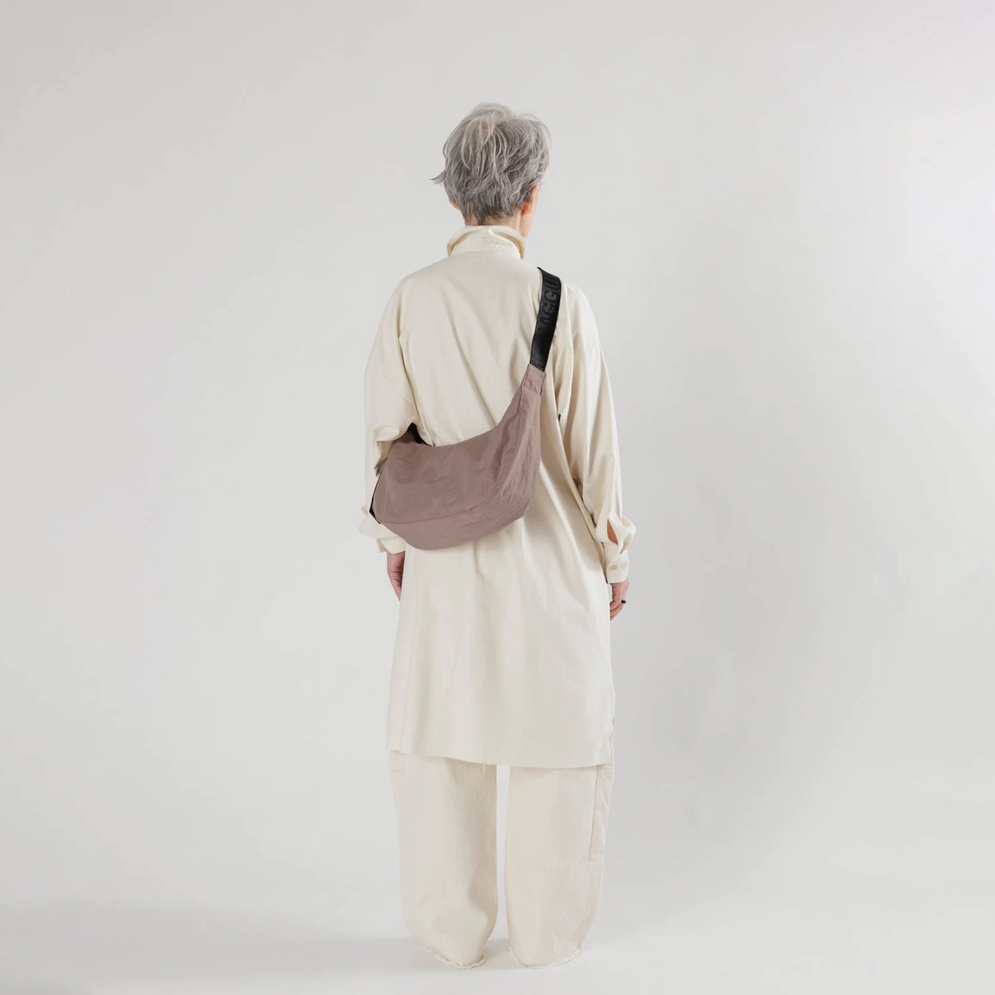 A taupe nylon crescent shaped bag with an adjustable black strap that reads, &quot;BAGGU&quot; in black and a single zipper.