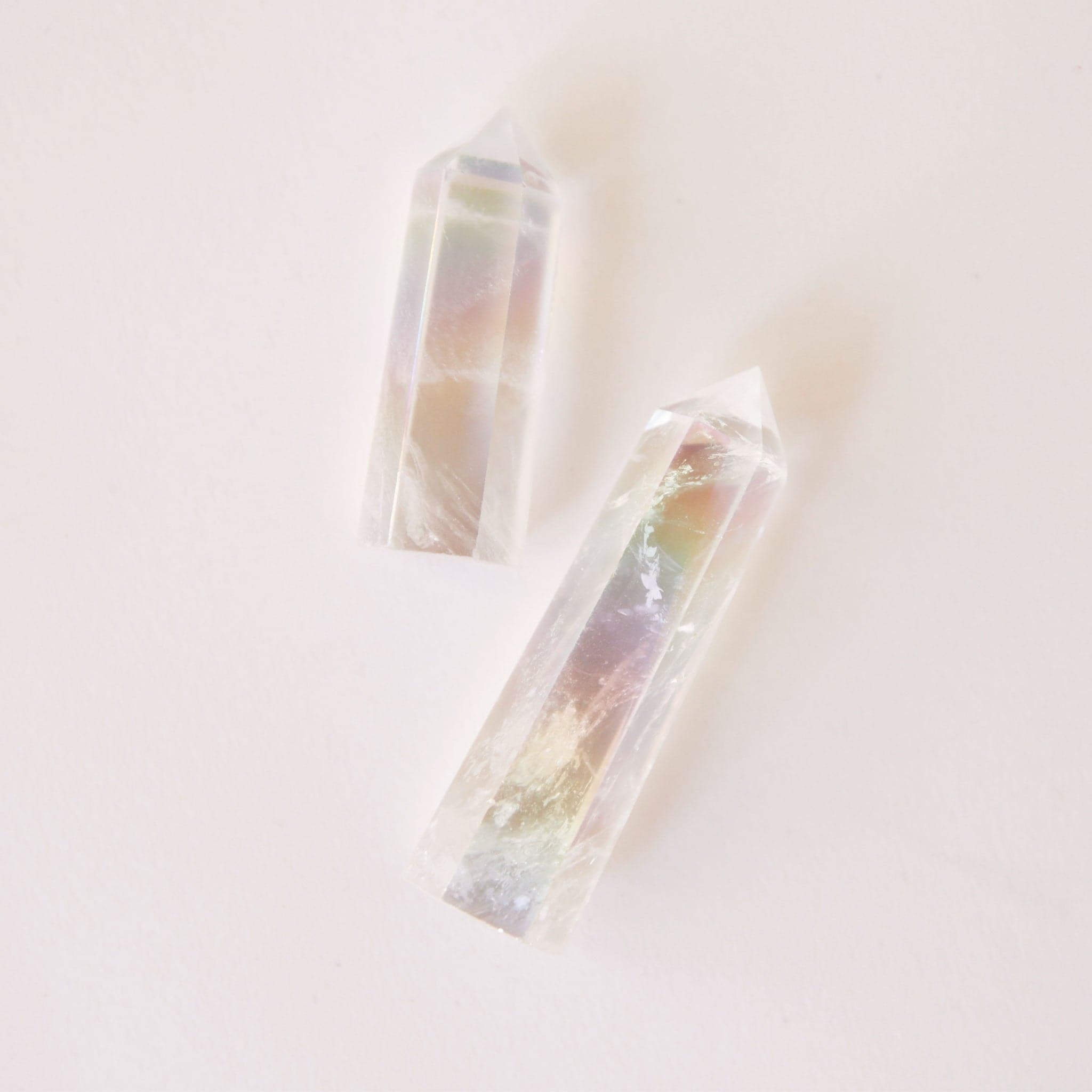 A photo of two aura clear quartz crystals, translucent with white accents and rainbow hues on the inside. The two sizes represent the two sizes available for purchase. Each crystal is sold separately. These aura clear quartz crystals are in the shape of a point and are flat on the bottom to allow them to be freestanding.