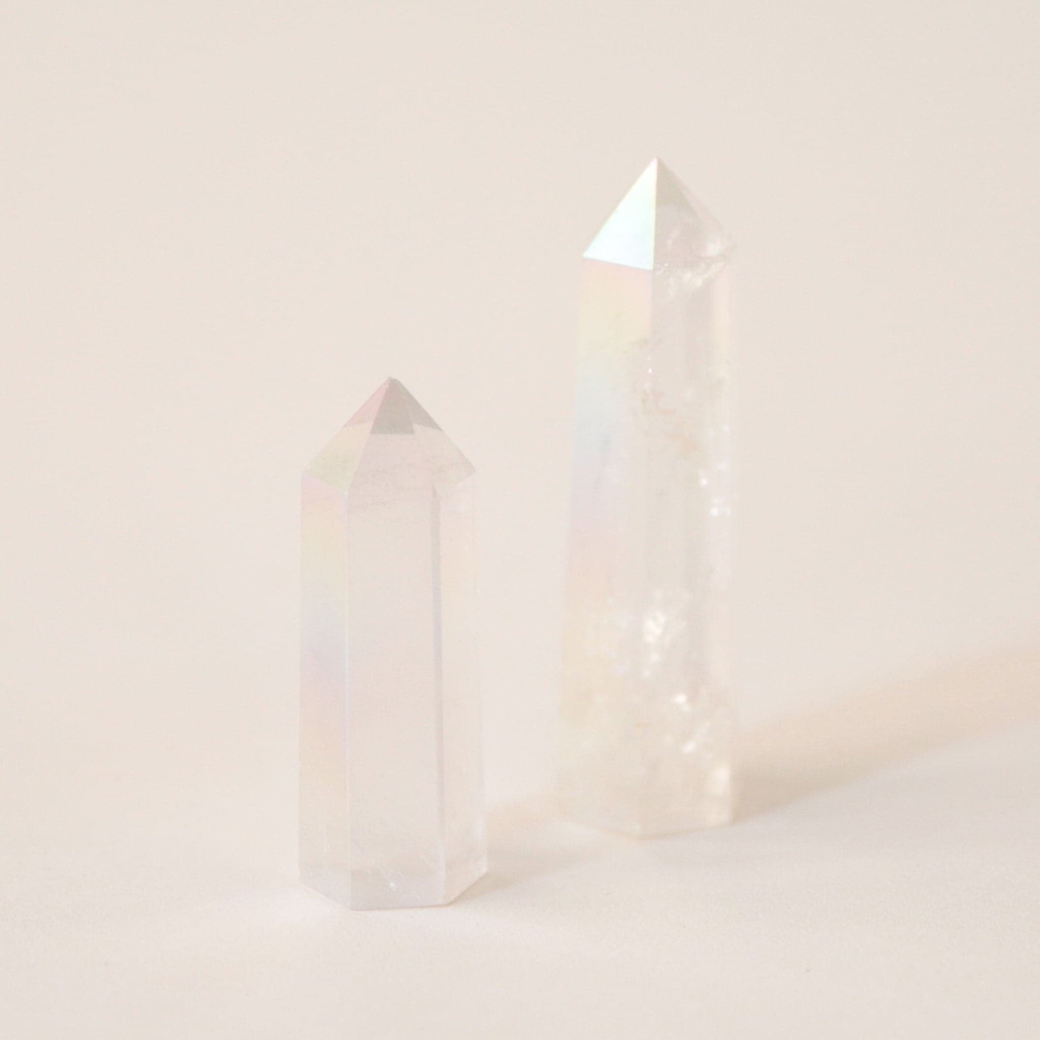 A photo of two aura clear quartz crystals, translucent with white accents and rainbow hues on the inside. The two sizes represent the two sizes available for purchase. Each crystal is sold separately. These aura clear quartz crystals are in the shape of a point and are flat on the bottom to allow them to be freestanding.