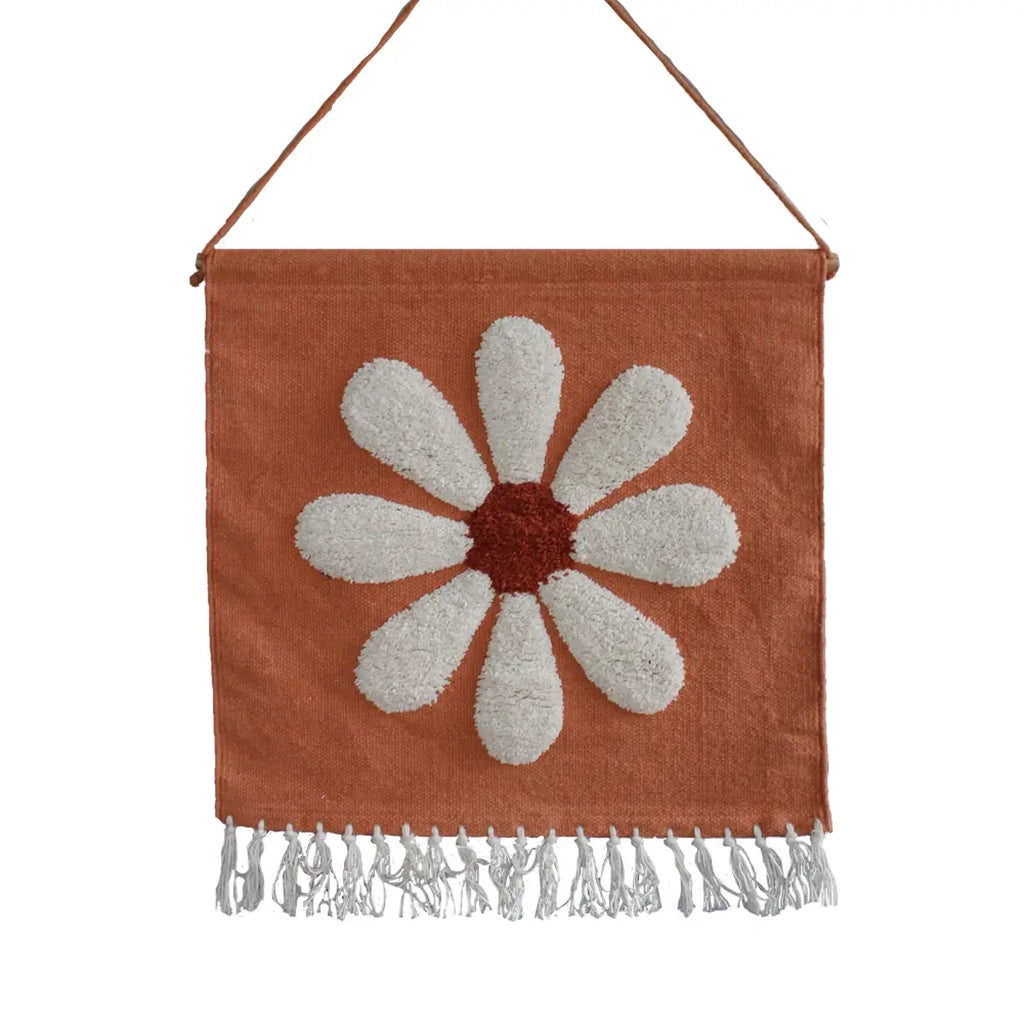 A terracotta hanging canvas with cream tassel detailing along the bottom edge and a white and rust daisy in the center.