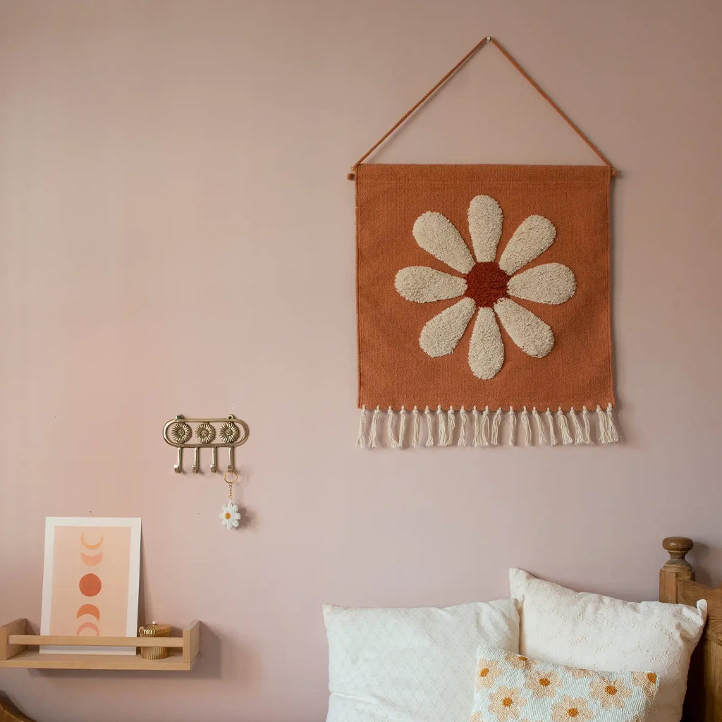 A terracotta hanging canvas with cream tassel detailing along the bottom edge and a white and rust daisy in the center. 