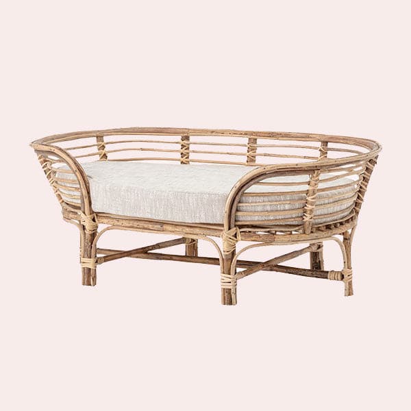 In front of a soft pink background is a rattan dog bed. The dog bed is an oval shape with six rattan legs on the bottom. Inside the bed is a light gray bed cushion. 