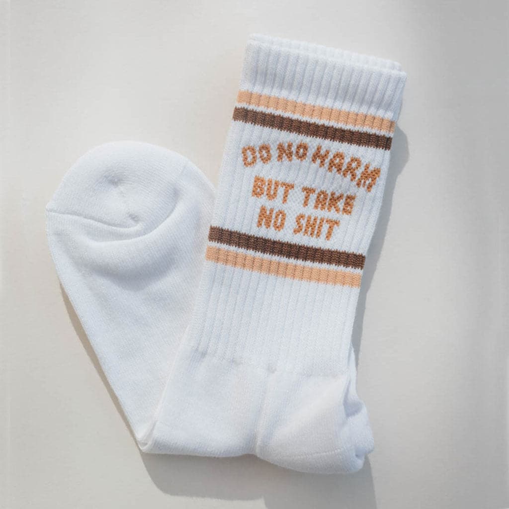 Pair of ribbed white tube socks that read &#39;do no harm but take no shit&#39; in burnt orange lettering. Two brown stripes fall above and below the text giving them a retro look.