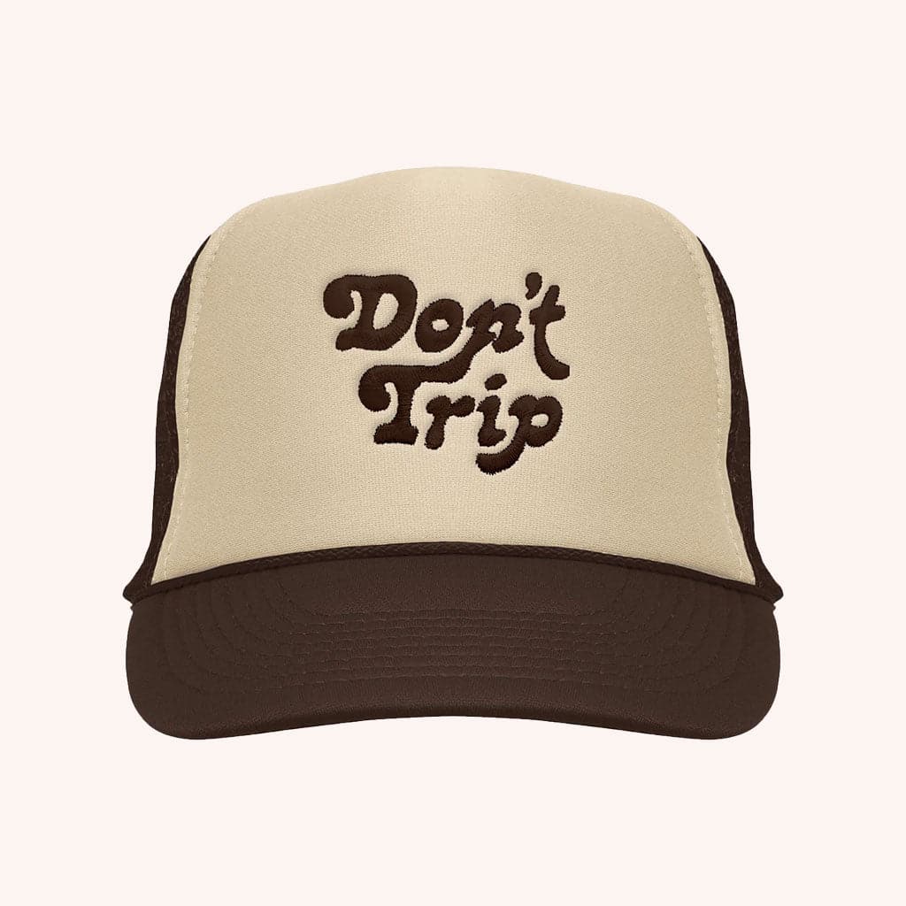 On a white background is a tan and dark brown trucker hat with text on the front that reads, &quot;Don&#39;t Trip&quot; in brown letters. 