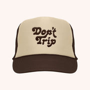 On a white background is a tan and dark brown trucker hat with text on the front that reads, "Don't Trip" in brown letters. 