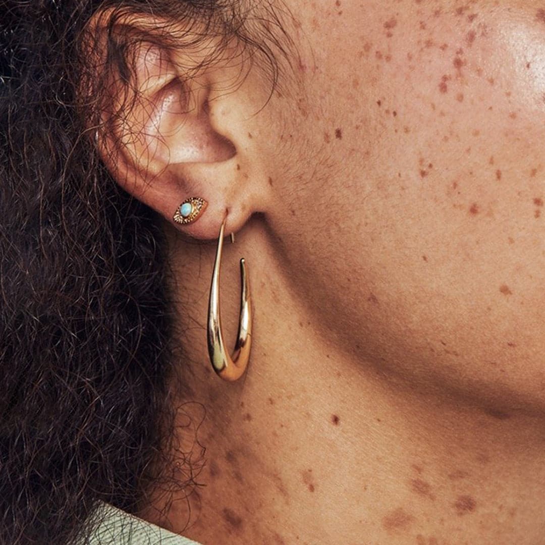 A gold oval shaped hoop earring with a thicker center piece compared to the sides of the hoops, modeled on an ear with other stud earrings in a stack.