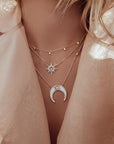 In this picture is a close up of a woman’s chest. She is wearing a pink, shimmery collared top. Around her neck are three gold necklaces. The middle necklace has a gold chain with a round moonstone with a gold border in the middle. There are sun beams extending from the moonstone. The sunbeams go back and forth from long to short