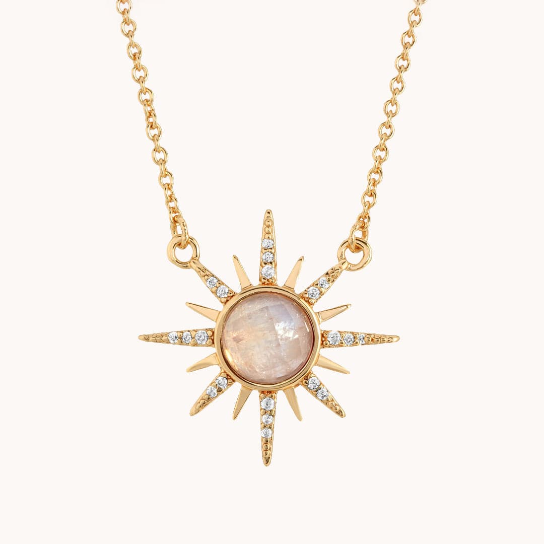 In front of a soft pink background is a gold chain. On the end of the chain is a round, moonstone with a gold border. There are sun beams extending from the moonstone. The sunbeams go back and forth from long to short. On the long sunbeams there are little clear crystals. 