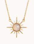 In front of a soft pink background is a gold chain. On the end of the chain is a round, moonstone with a gold border. There are sun beams extending from the moonstone. The sunbeams go back and forth from long to short. On the long sunbeams there are little clear crystals. 