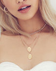 Woman modeling two layered Aura Pendant Necklaces. Gold oval pendant with set clear crystal gemstone in center and gold indentations like sun rays in the gold pendant.