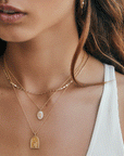This is a picture of a woman with long brown hair. She is wearing a white tank top. Around her neck is three gold chain necklaces. The longest necklace has a gold pendant on the bottom. The pendant has an arched top and straight across bottom. On the front is gold waves and two gold palm trees. Above the trees are gold stars.