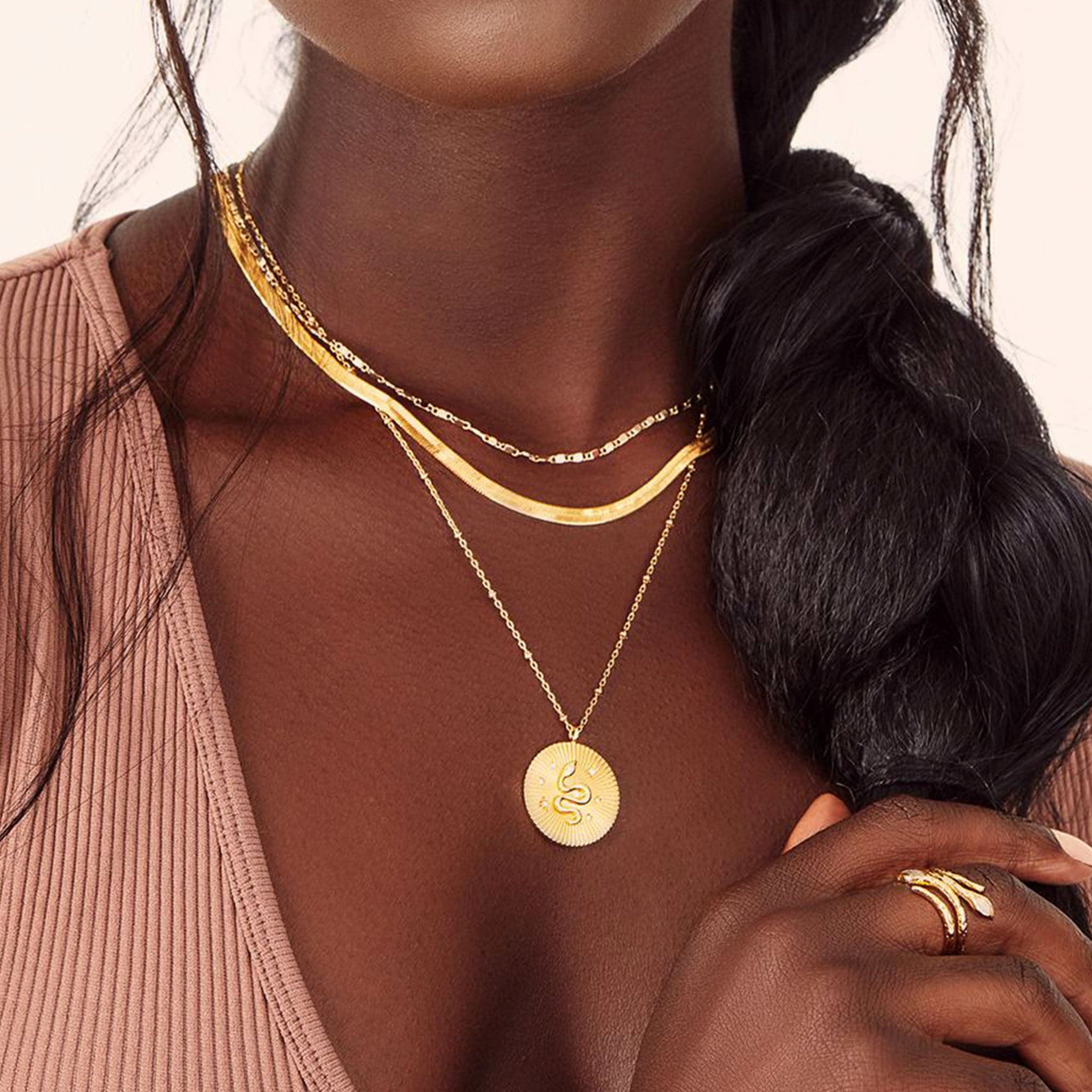 On a white background is a gold necklace with a round pendant in the center that has a snake detail with CZ stones placed around it worn on a model with other necklaces. 
