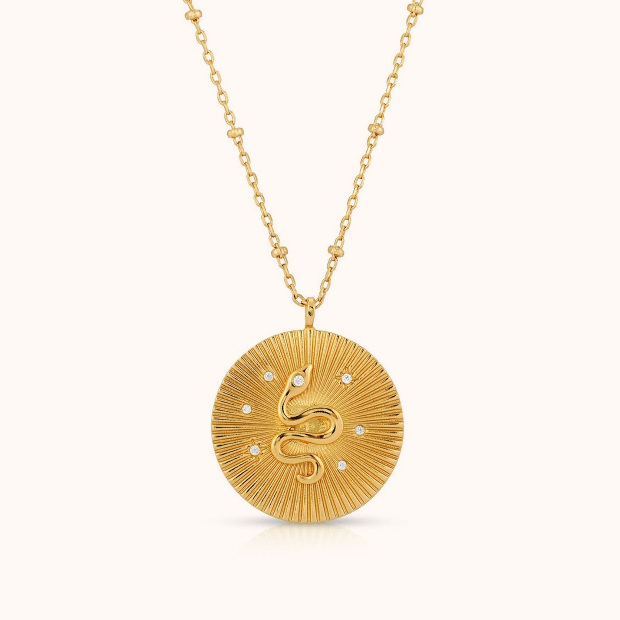 On a white background is a gold necklace with a round pendant in the center that has a snake detail with CZ stones placed around it. 