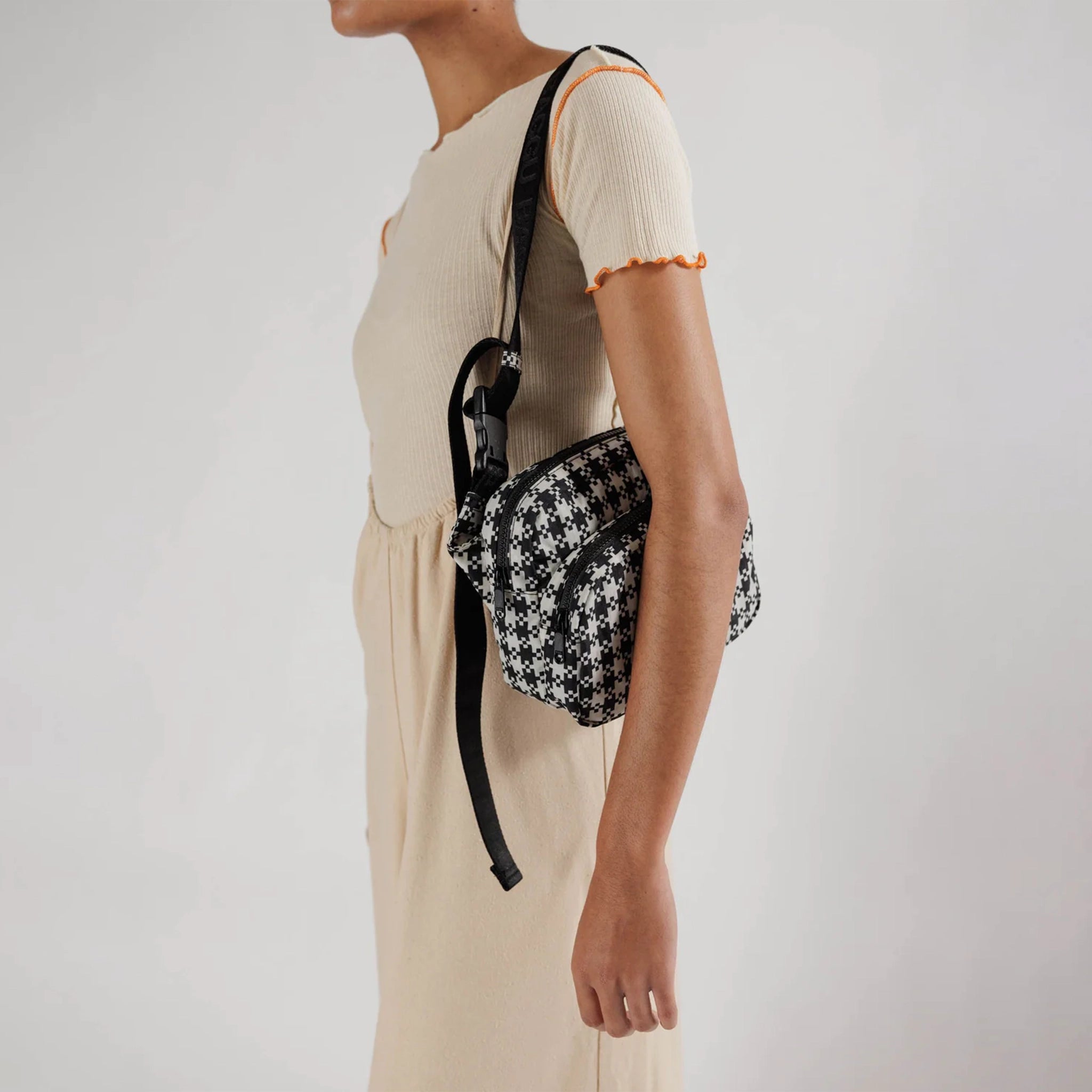 A black and white gingham nylon fanny pack with two zipper pockets and a black adjustable strap modeled over someones shoulder and chest.