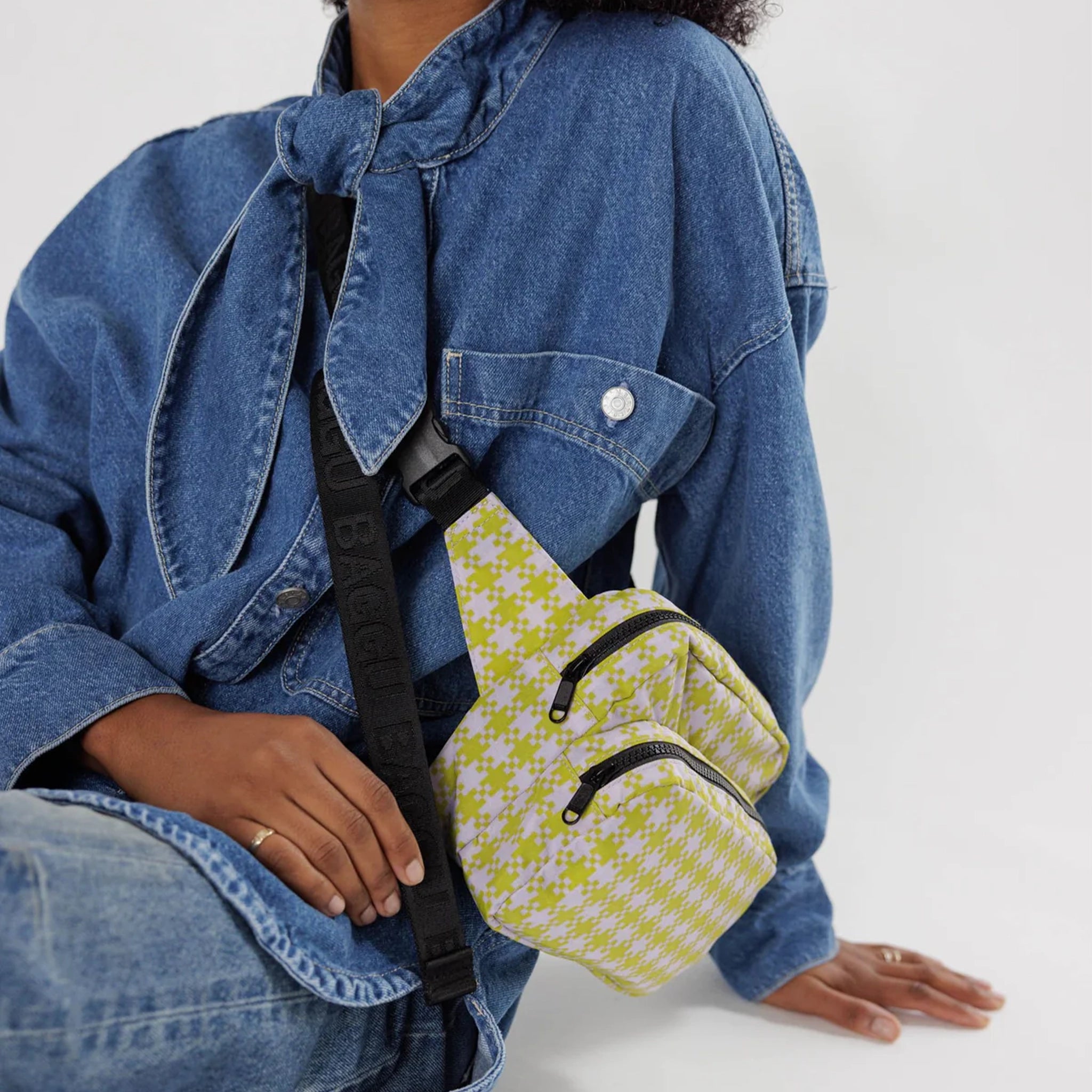 A light pink and lime green gingham nylon fanny pack with two zipper pockets and a black adjustable strap modeled over someones shoulder and chest.