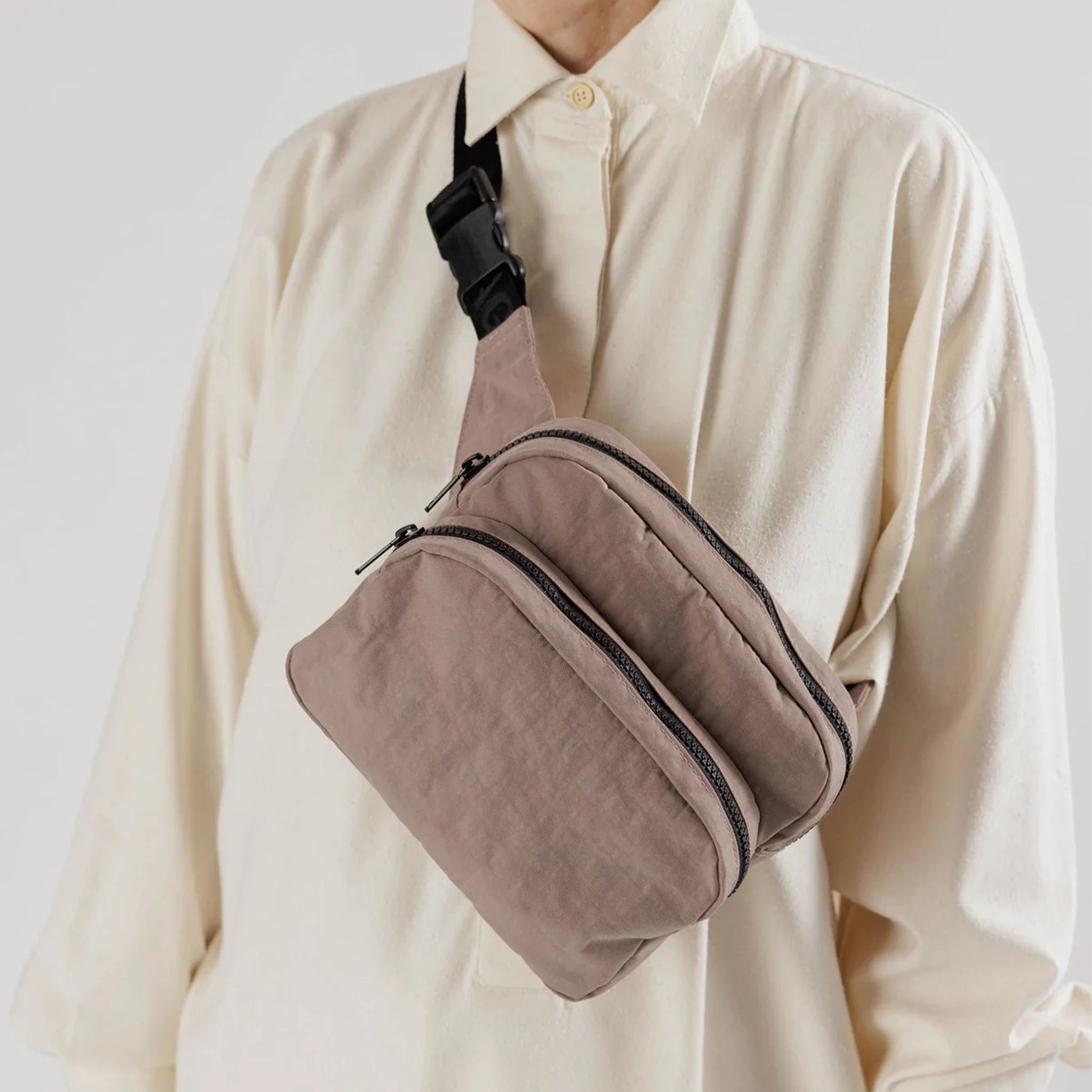  A taupe nylon fanny pack with two zipper pockets and a black adjustable strap modeled over someones shoulder and chest.
