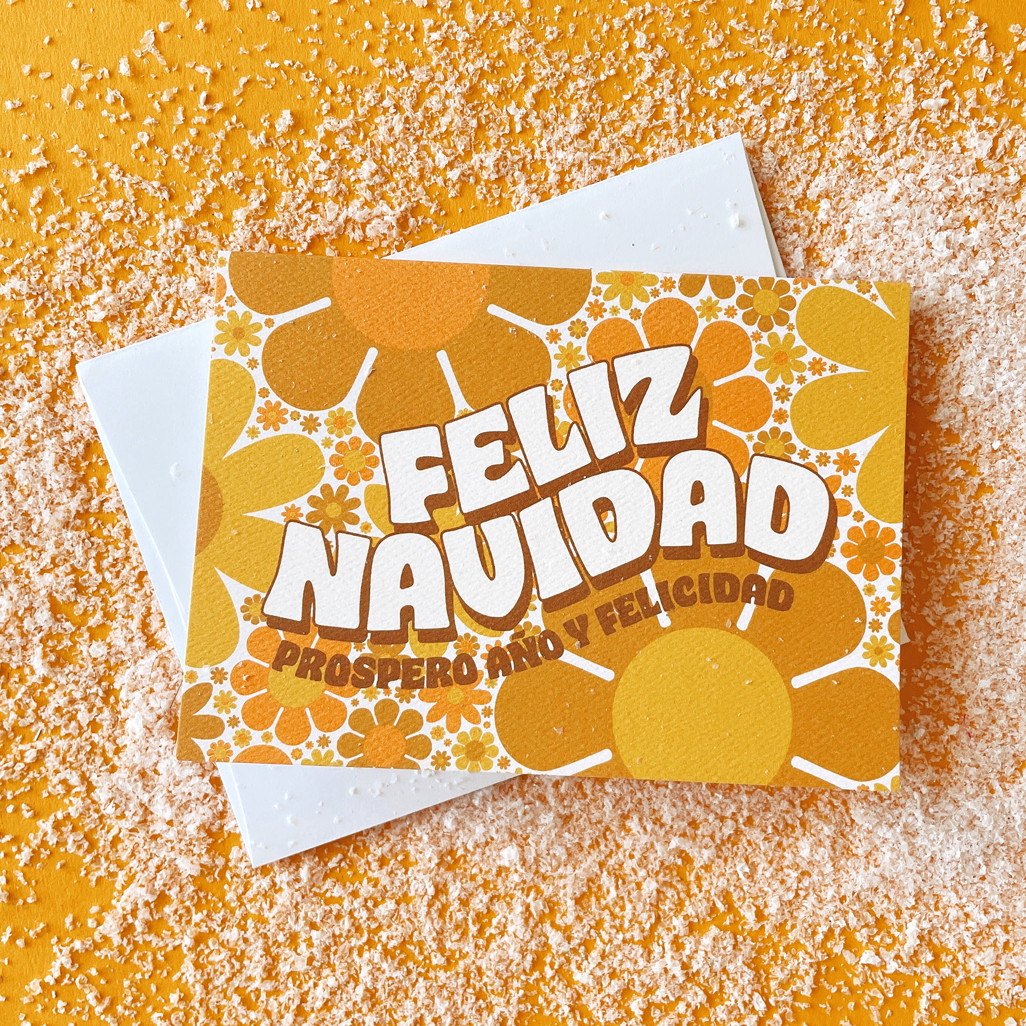 Greeting card filled with yellow and orange retro flower print. The card reads &#39;Feliz Navidad&#39; in white curved bubble letters. Below reads &#39;prosper año y felicidad&#39; in rust colored lettering. The card is accompanied by a solid white envelope.