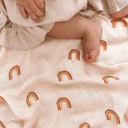 Neutral toned swaddle with warm rainbow design displayed under a baby with sweet button toes and a tan ruffly skirt.