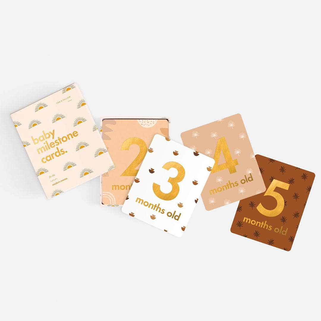 Ivory book with gold sun illustration and gold foil text "Baby Milestone Cards - For the modern mama." With four cards pulled out, "2 months old," "3 months old," "4 months old," "5 months old."