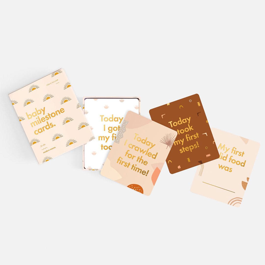 Ivory book with gold sun illustration and gold foil text &quot;Baby Milestone Cards - For the modern mama.&quot; With four cards pulled out, &quot;Today I got my first tooth,&quot; &quot;Today I crawled for the first time,&quot; &quot;Today I took my first steps,&quot; &quot;My first solid food was.&quot;