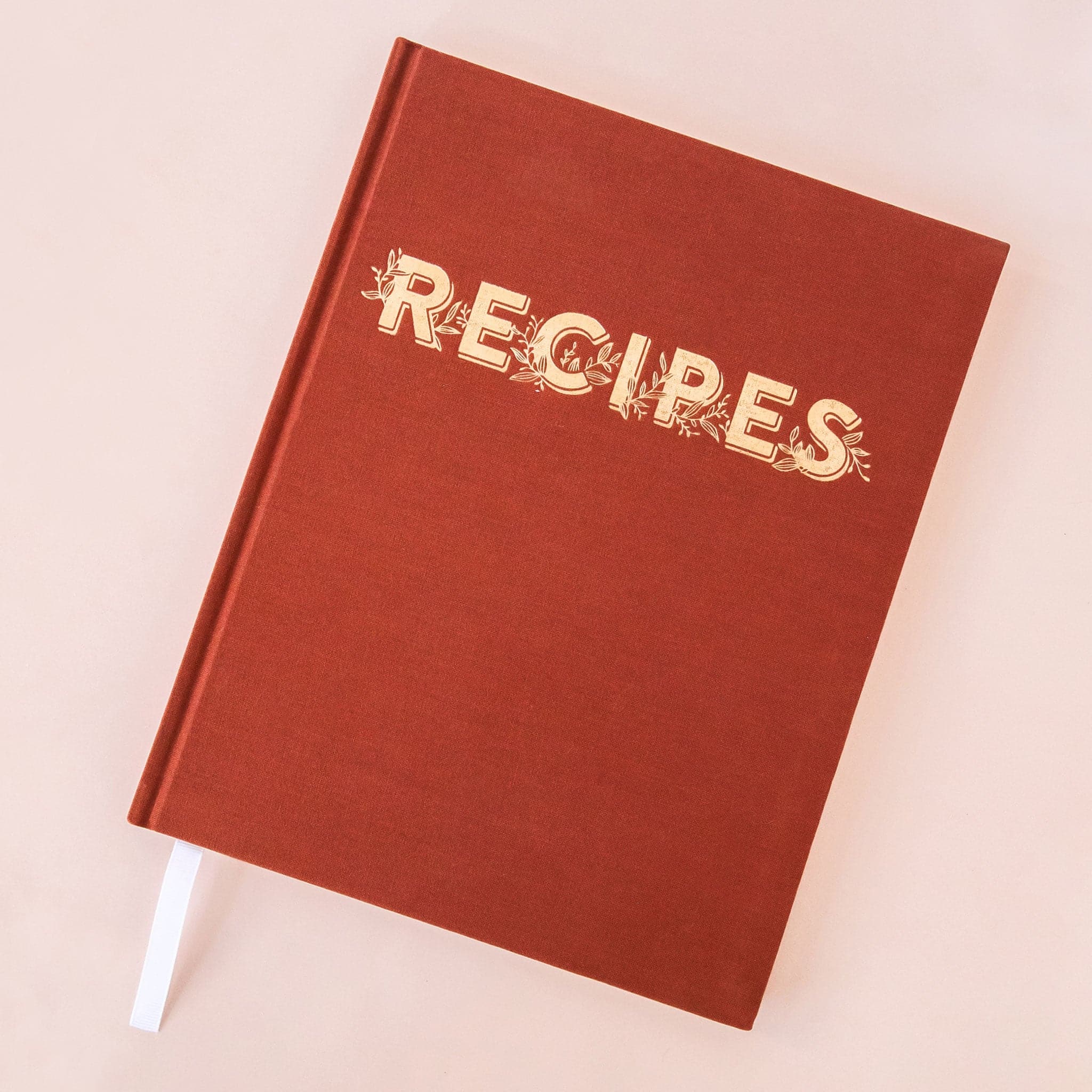 In front of a pink background is the cover of a red book. Towards the top is gold text that reads ‘Recipes.’ Intertwined with the letters is gold leaves. Sticking out of the bottom left corner is a white bookmark.