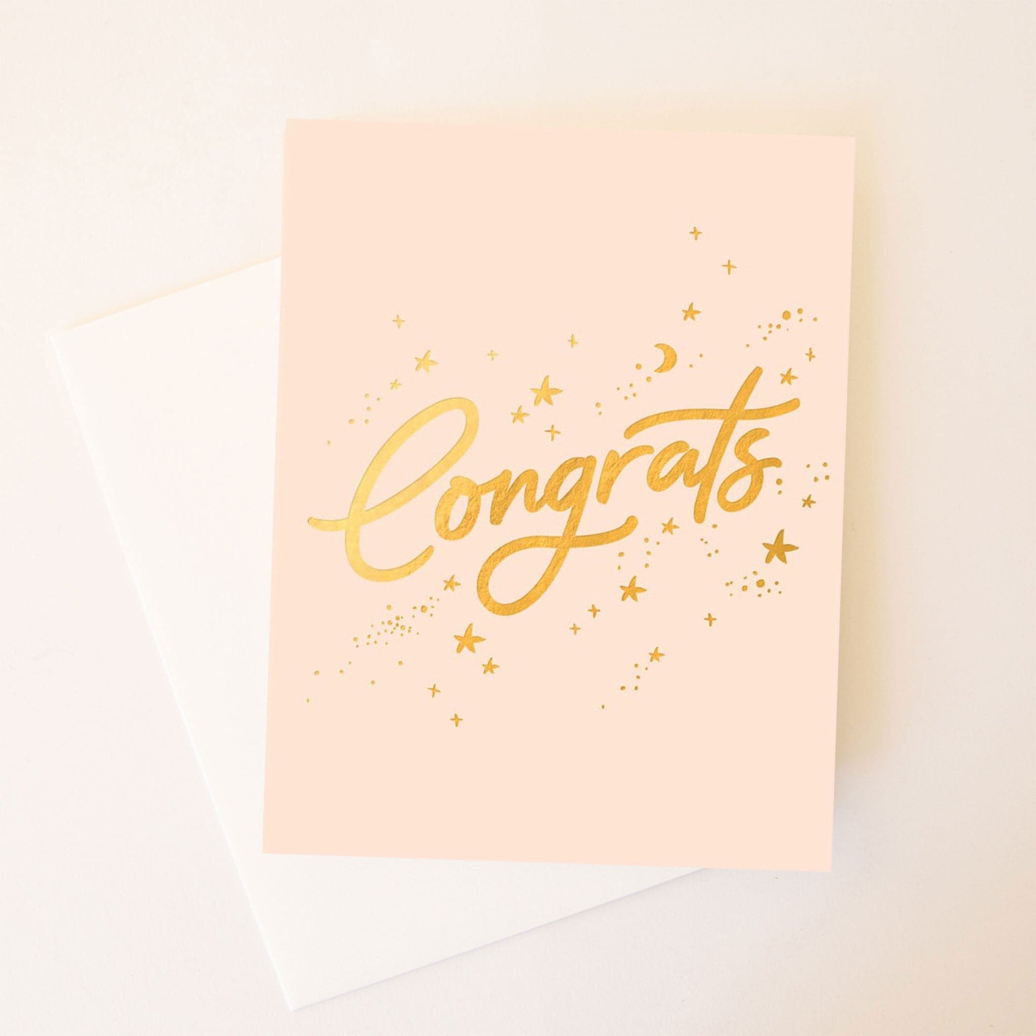 A light pink card with gold cursive writing on the front that says, "Congrats" along with small stars and moons around the word.