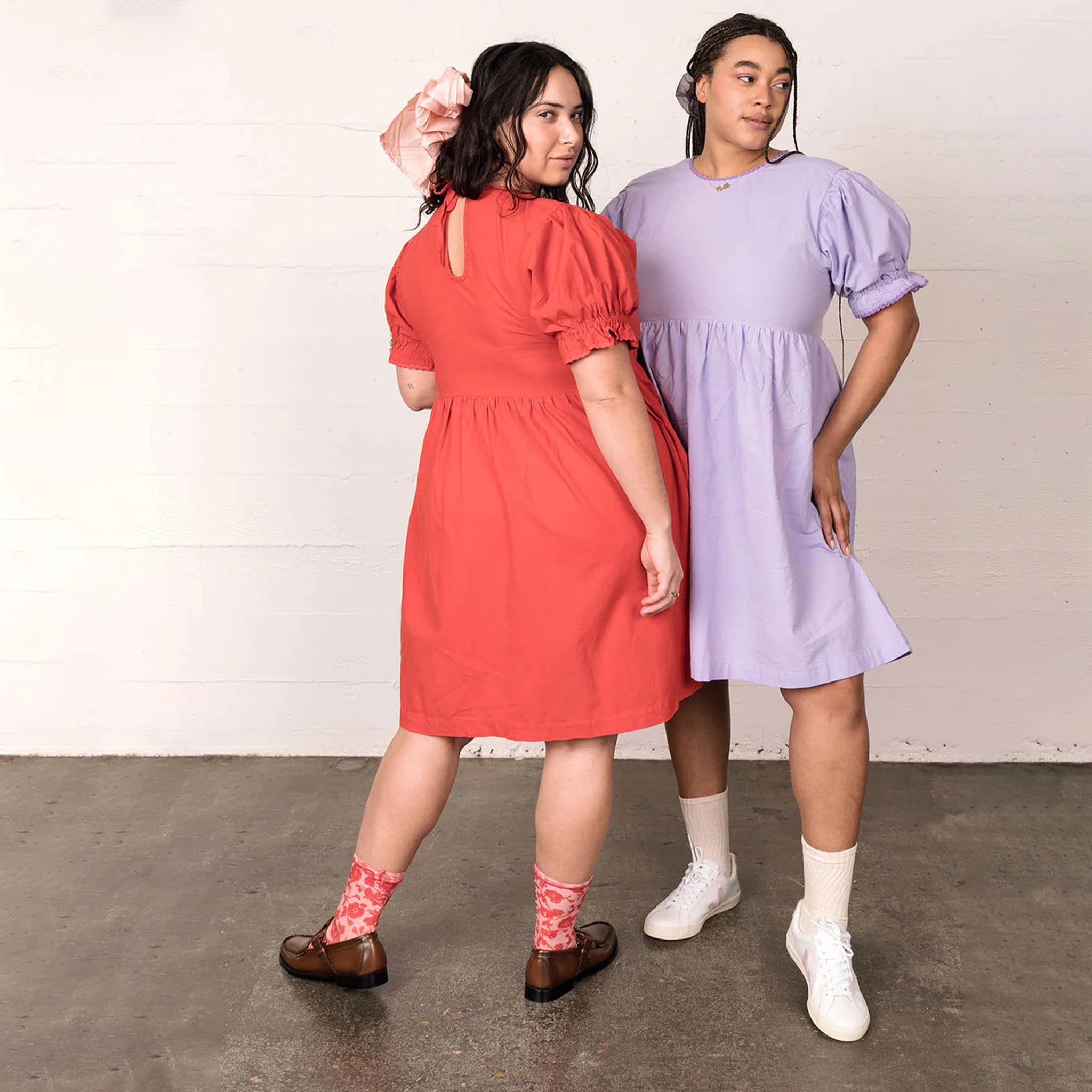 In front of a cream wall is a model wearing a lavender baby doll style dress with puffy sleeve details next to another model wearing the same dress in red. 