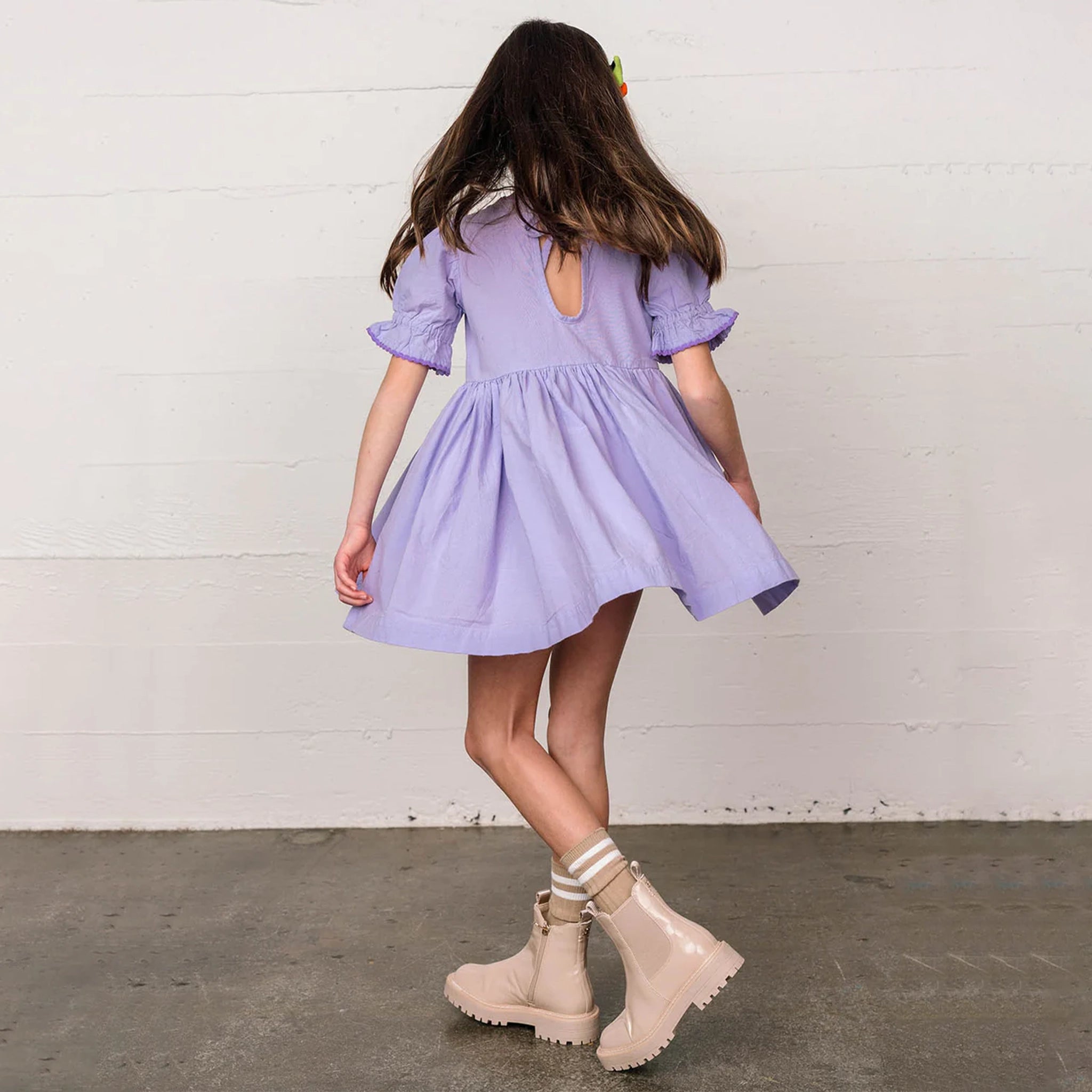 In front of a cream wall is a children's model wearing a lavender baby doll style dress with puff sleeves and a flowy skirt that hits right above the knee.