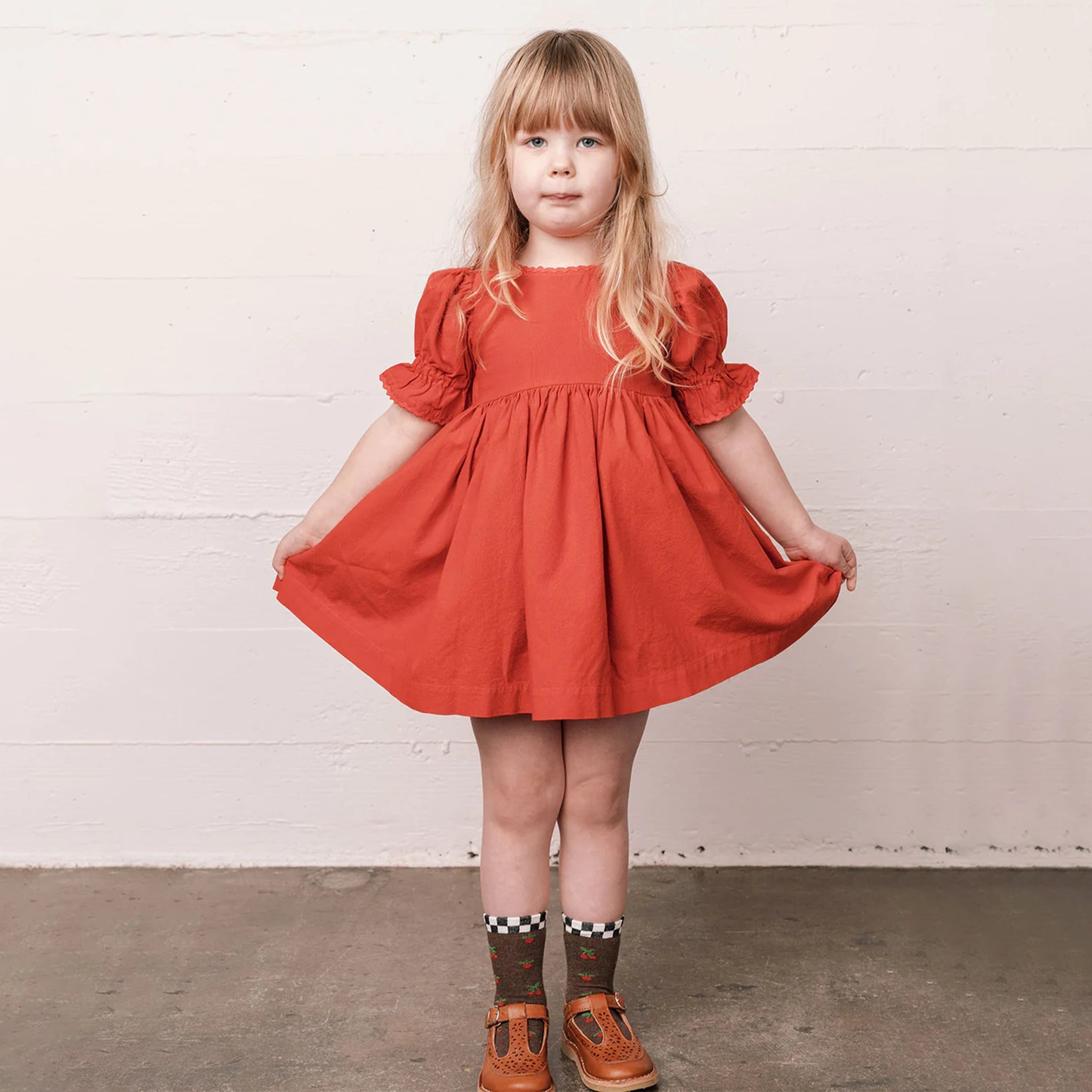 In front of a cream wall is a children's model wearing a red baby doll style dress with puffy sleeve details.