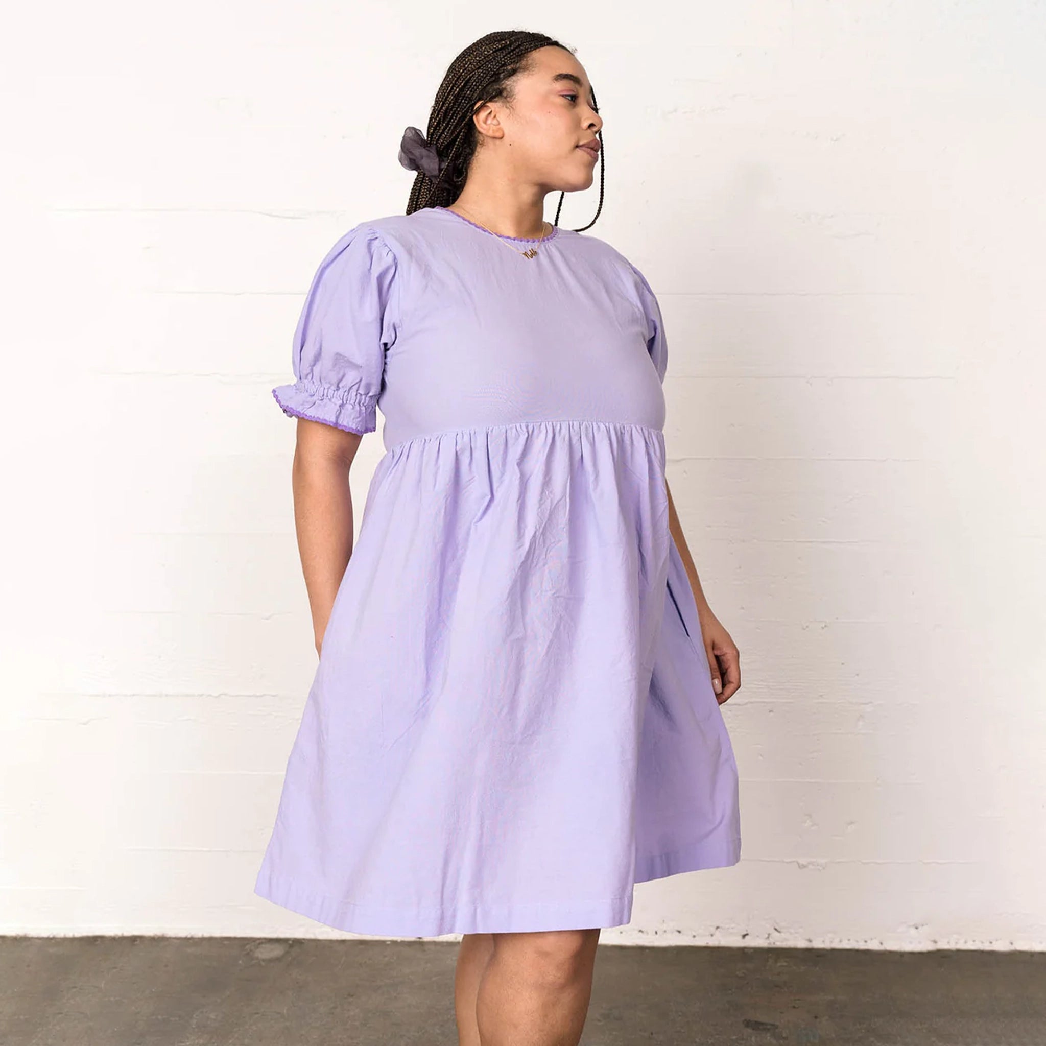 In front of a cream wall is a model wearing a lavender baby doll style dress with puffy sleeve details.