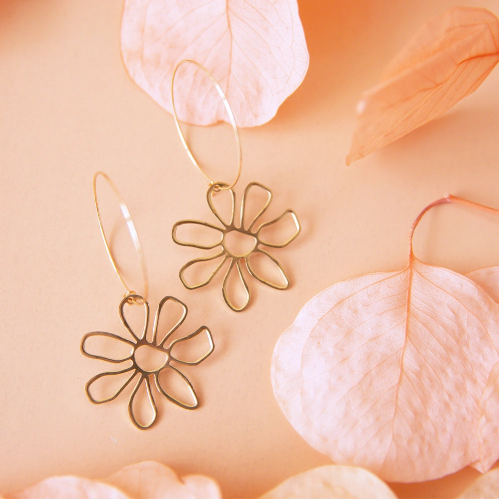 On a peachy background is a pair of gold hoop earrings with a thin flower charm hanging from the bottom of the hoop. The earrings are staged next to pink dried floral petals. 