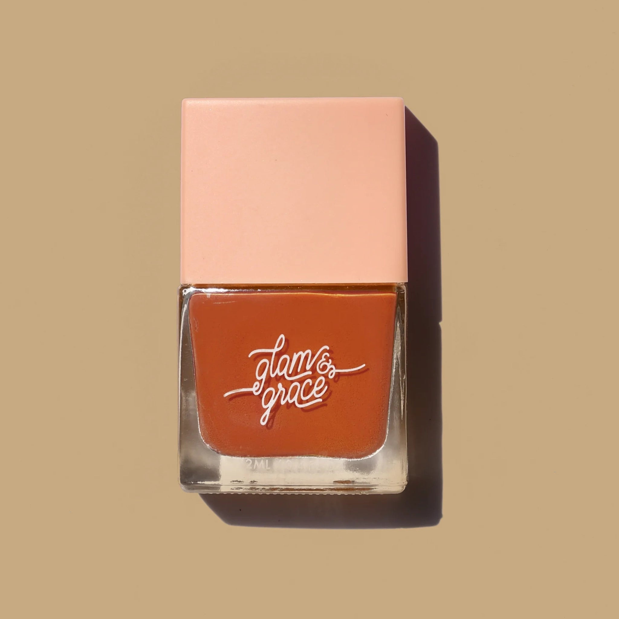 A burnt orange nail polish in a square glass jar with a salmon pink lid along with white cursive text on the front that reads, "glam & grace".