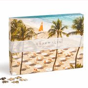 A boxed puzzle with a seaside aerial photograph from Gray’s best-selling collection, À la Plage, featuring a beach with umbrellas, sun chairs and palm trees.