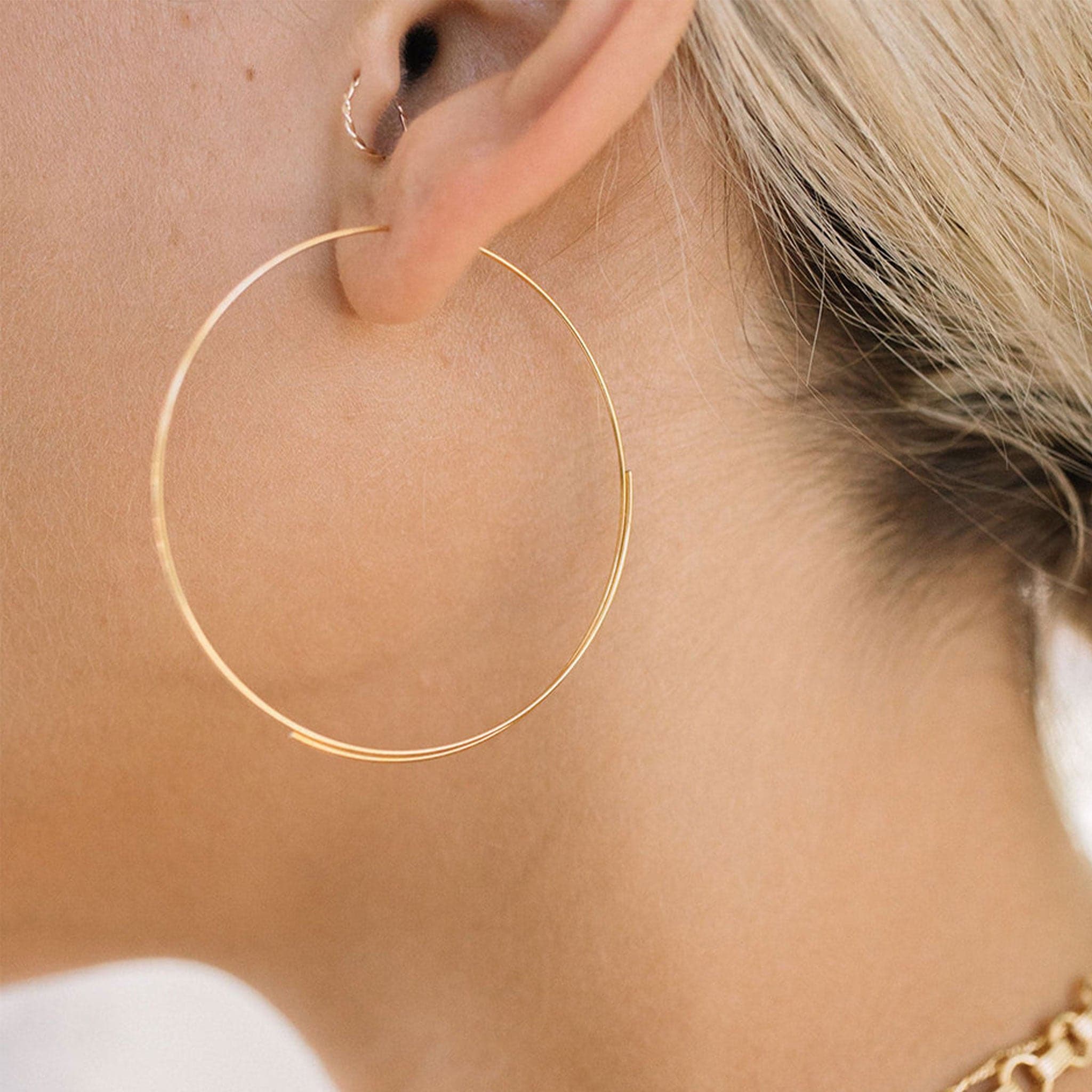 A model wearing the Hadley Hoops which are thin larger gold hoop earrings. 