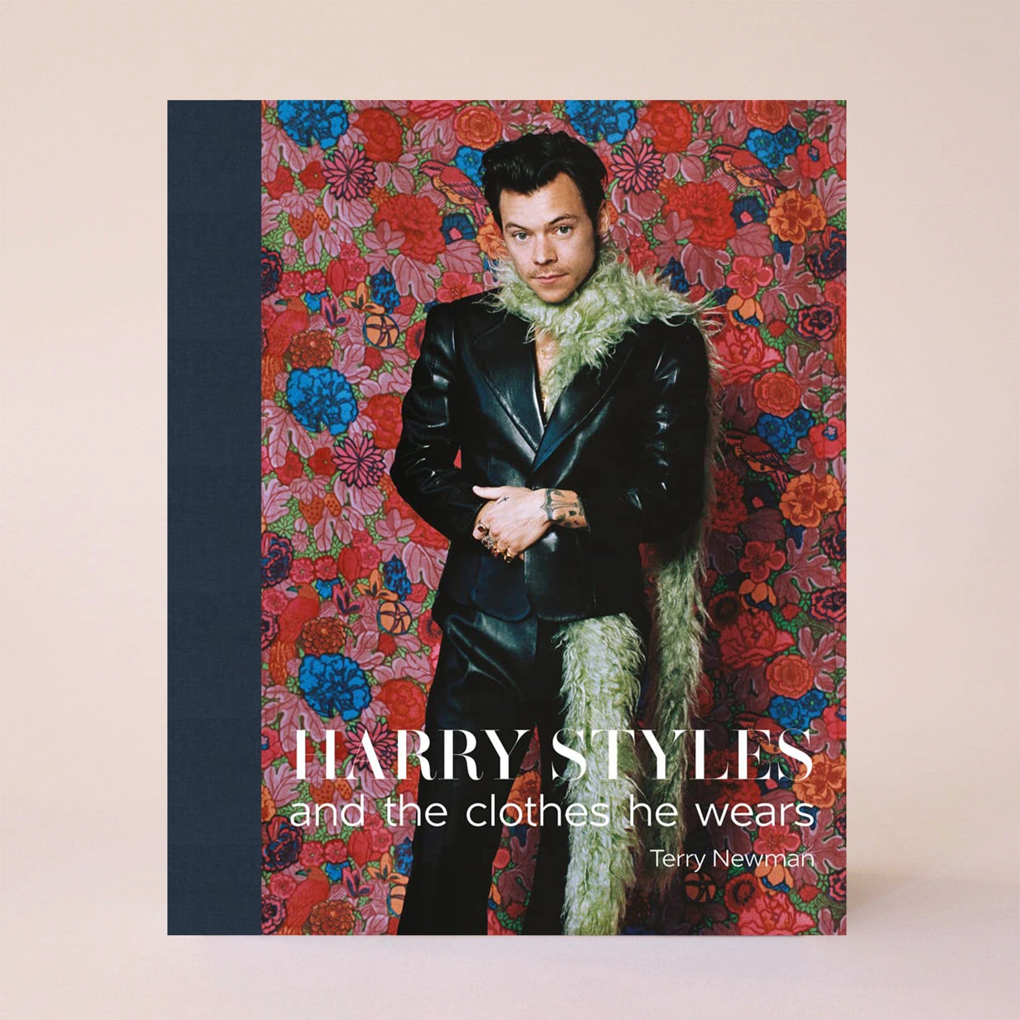 On a cream background is a book cover with a navy blue spine and a red, pink and blue floral design behind the iconic Harry Styles in a black suit and a green boa along with text on the bottom that reads, &quot;Harry Styles and the clothes he wears&quot; in white letters. 