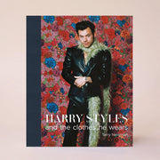 On a cream background is a book cover with a navy blue spine and a red, pink and blue floral design behind the iconic Harry Styles in a black suit and a green boa along with text on the bottom that reads, "Harry Styles and the clothes he wears" in white letters. 