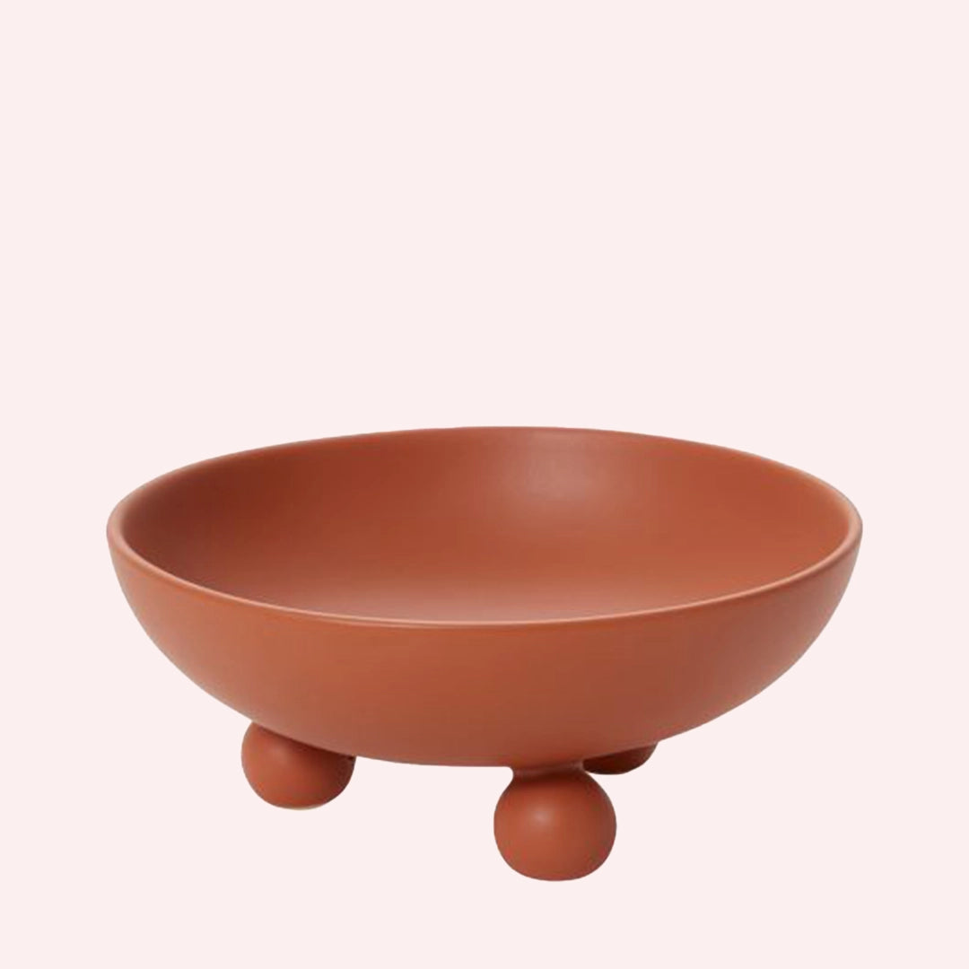 A ceramic wide mouth bowl in a matte terracotta shade with three sphere feet.