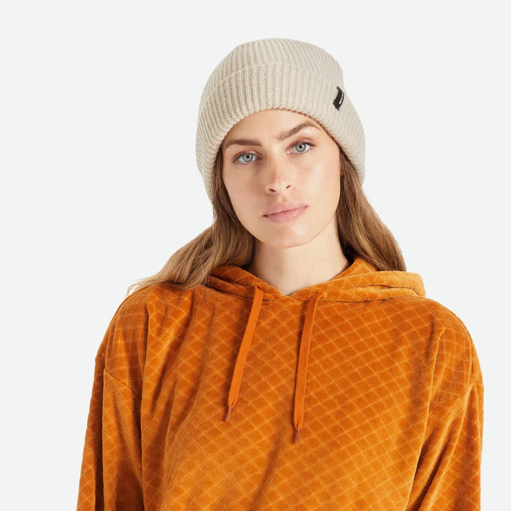 A knit ribbed beanie in a beige color with a folded brim and a black label on the front right left corner.