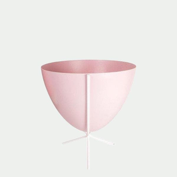 In front of white background is a pink bullet planter in a short white metal stand. The bullet planter is wide at the top and narrow at the bottom. The metal stand has three legs. 