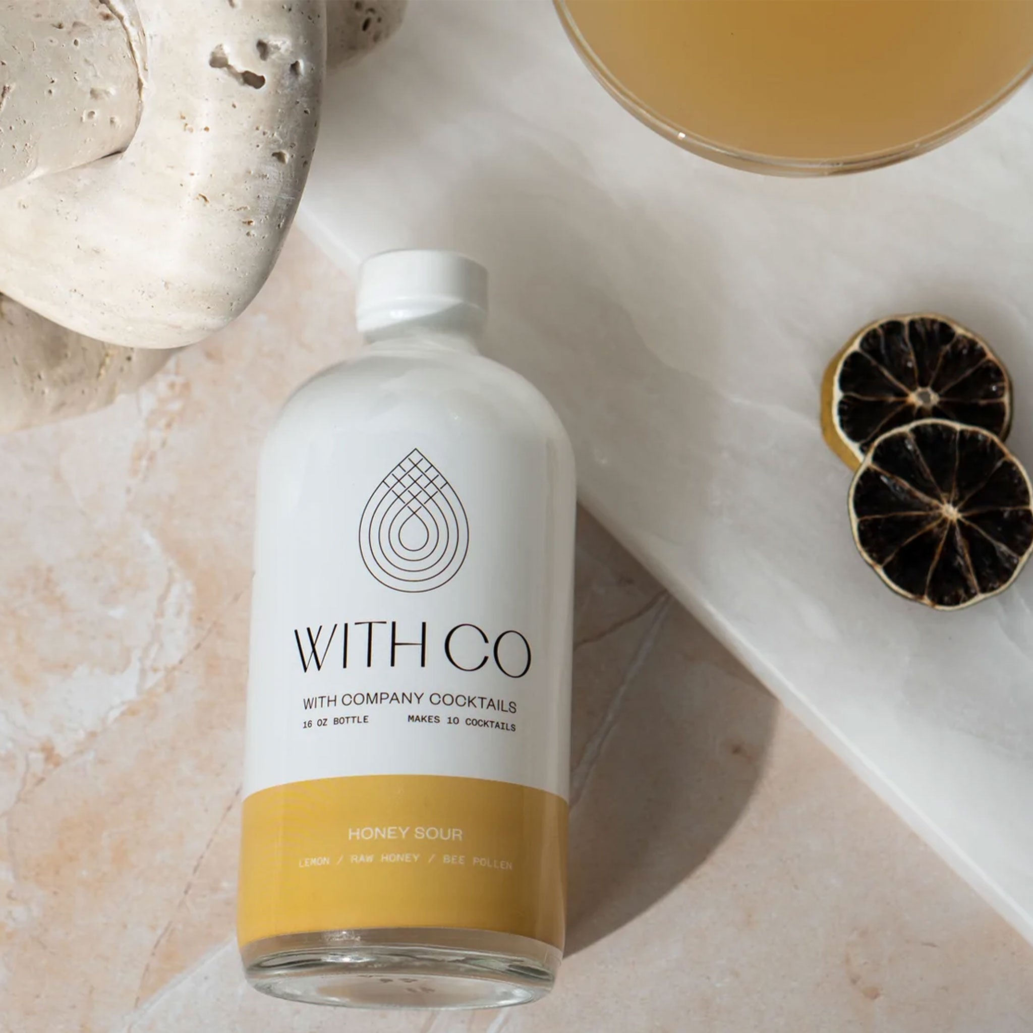 A white bottle of honey sour cocktail mix photographed on a neutral stone backdrop and two dried limes.