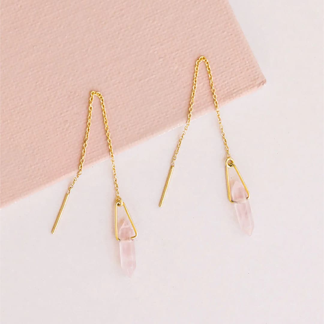 Pair of gold plated chain earrings. Each earring has a rose quartz stone positioned between a gold loop. Pair lays against a soft pink textured box and white counter. 