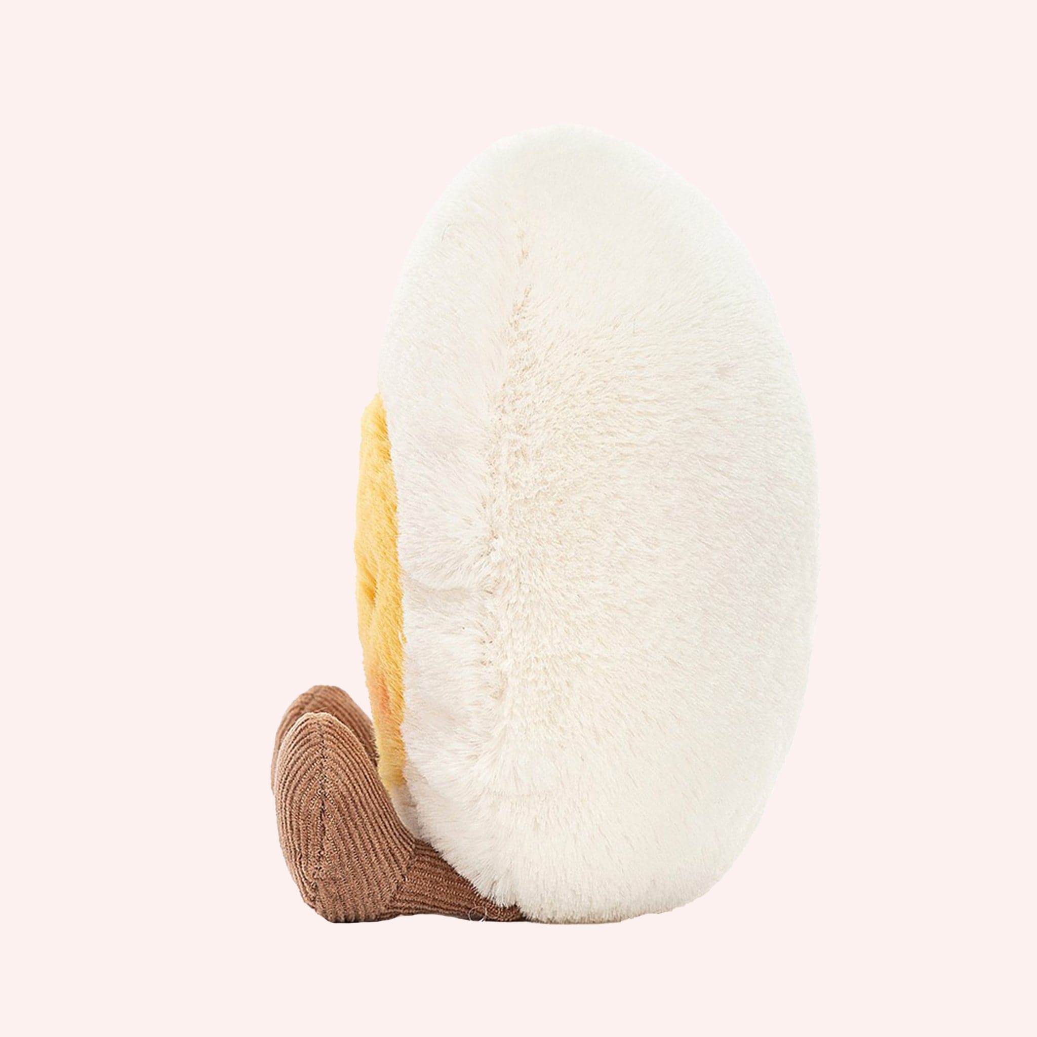 Side view of A soft stuffed animal in the shape of an egg with yellow yoke face and blushing smiley face, and brown floppy legs.