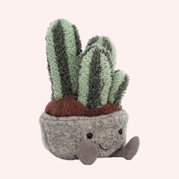In front of a soft pink background is a succulent stuffed animal. It is an oval shaped gray pot with two brown legs at the bottom. Inside the pot is brown with four different sized cacti. They are all dark green and light green vertical stripes. 