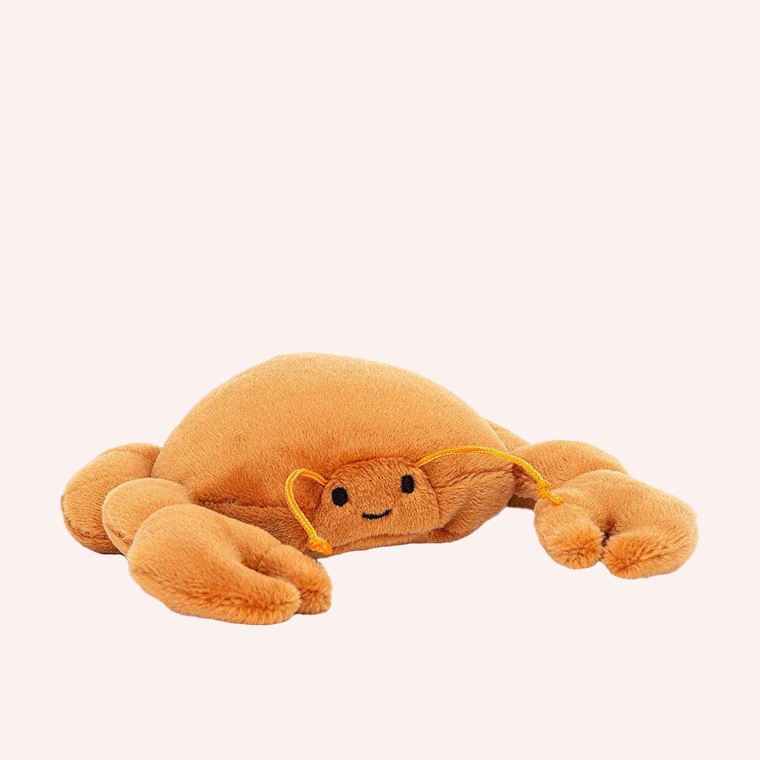 In front of a cream background is a stuffed orange crab laying on its stomach. It has two large claws in the front and two little claws in the back. On its face are two black eyes and a little black smile. He has two orange antennas above his eyes.