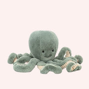 In front of a white background is a light blue stuffed octopus. On his head are two black eyes and a small black mouth. The bottom of his tentacles is cream colored. 