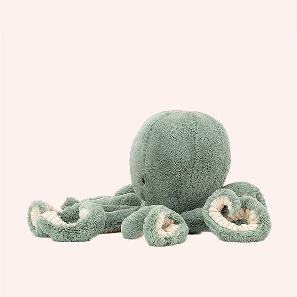 In front of a white background is a light blue stuffed octopus. On his head are two black eyes and a small black mouth. The bottom of his tentacles is cream colored. 