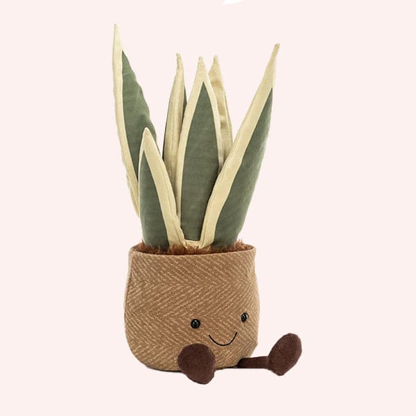 A soft suede stuffed animal in the shape of a potted snake plant, with spikey dark and light green leaves, and brown textured pot with smiley face and dark brown floppy legs.