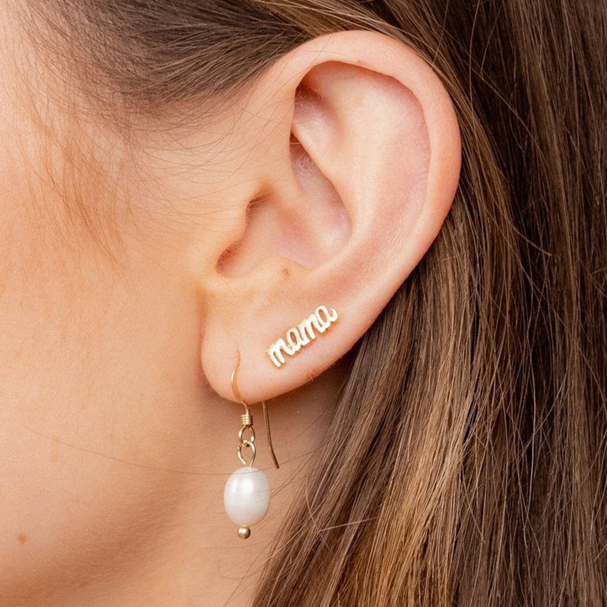 A model&#39;s ear wearing dainty gold stud earrings that say &quot;mama&quot;.