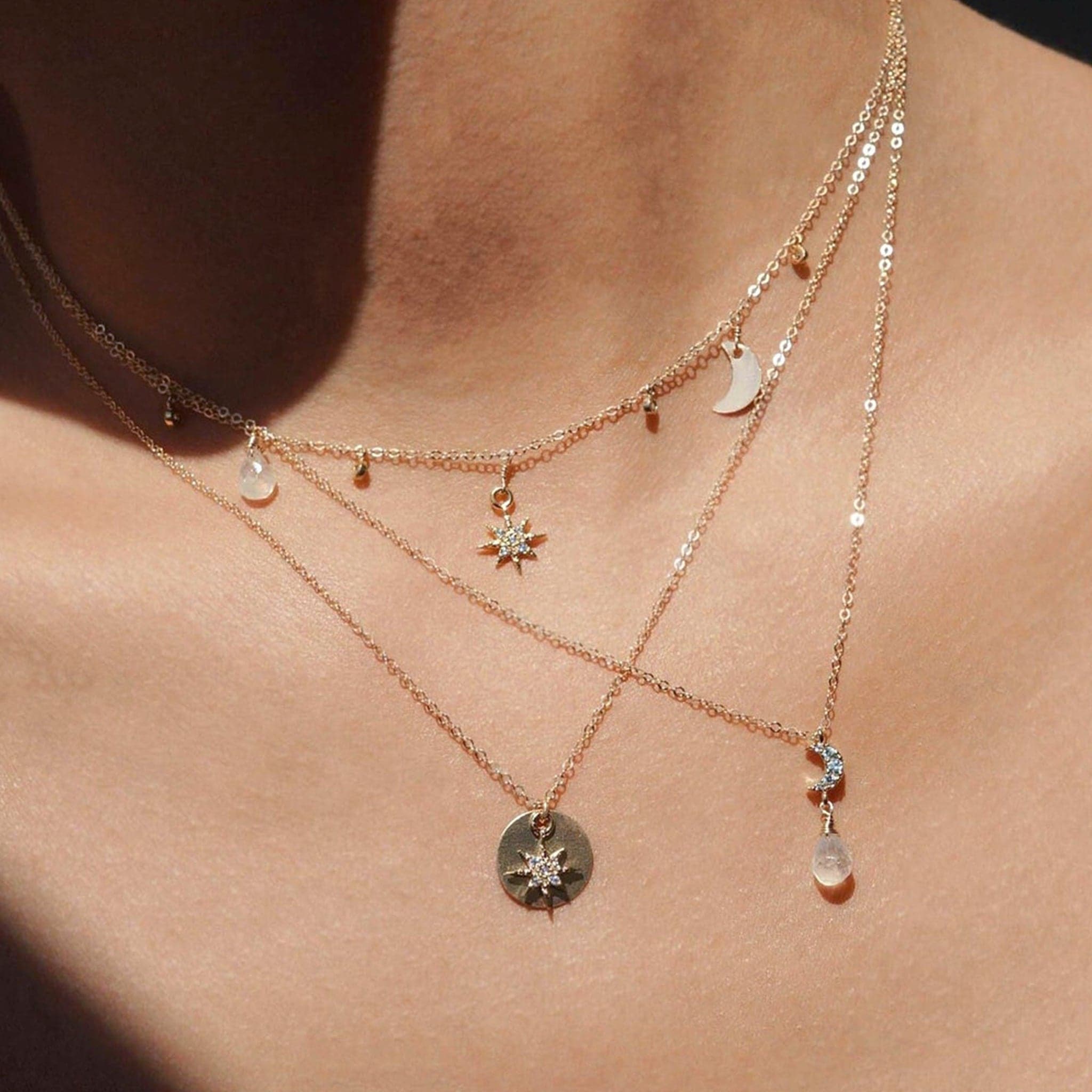Close up of three layered necklaces being worn. One is a gold choker with star, moon, and white gem charms. One necklace is a long gold necklace with silver gem moon with white gem drop. The adenna necklace is the third necklace. Long gold chain and circle pendant with crystal gemmed star charm.