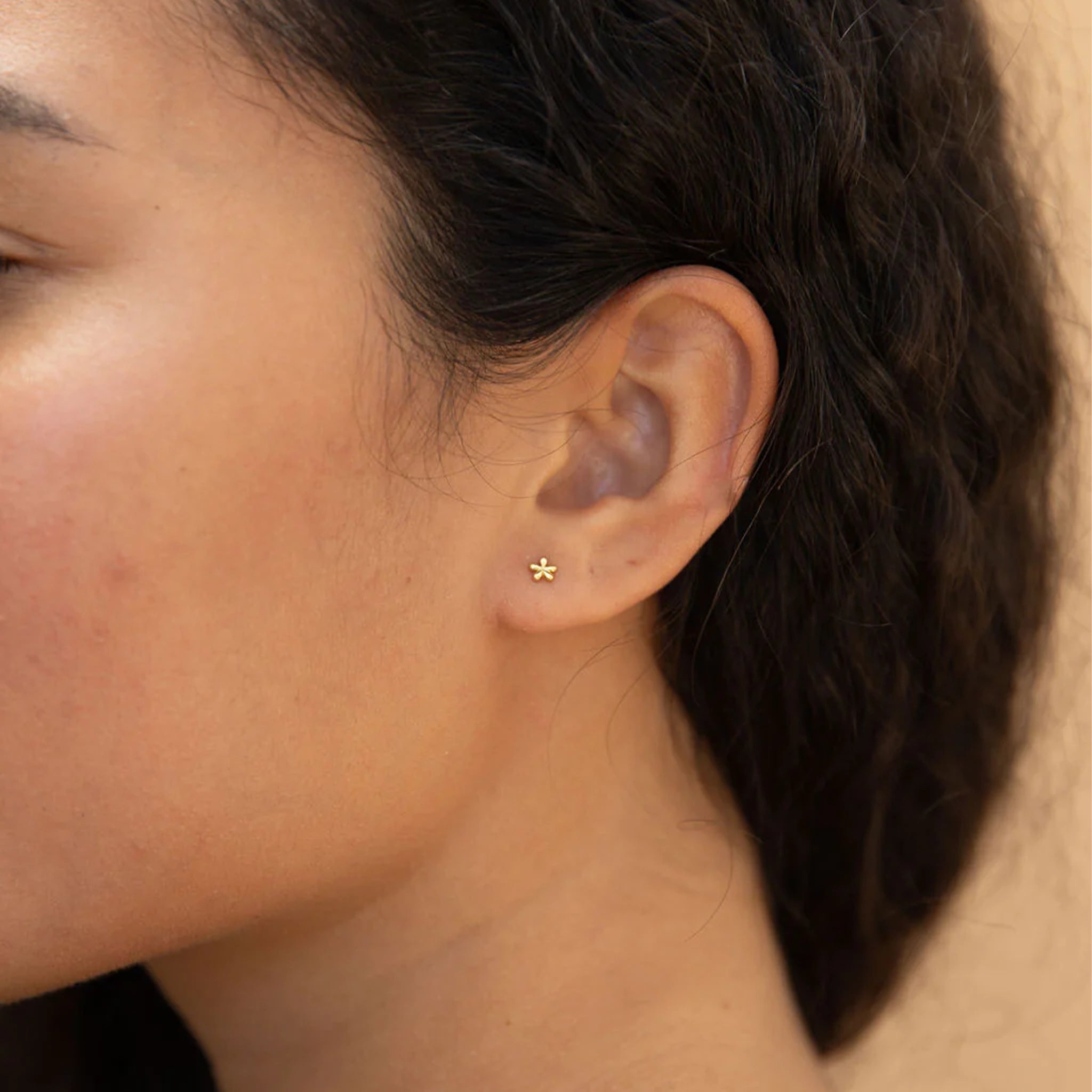 A pair of tiny gold daisy shaped stud earrings with a straight gold post worn here on a model's ear.
